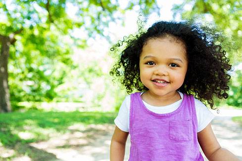 Cute African American little girl walks in the park on a sunny day. The wind is blowing her curly black hair. She has brown eyes and is wearing a purple tank top with a white t-shirt underneath. Grass and trees are blurred in the background. Copy space available.